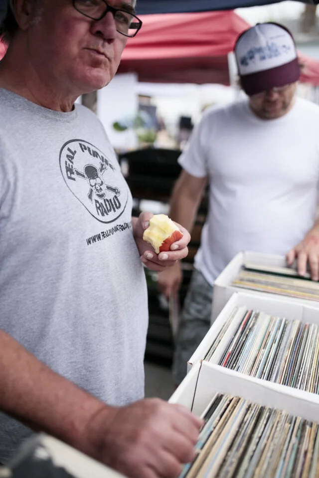 Two people browse vinyl records at an outdoor stall beneath a red canopy.