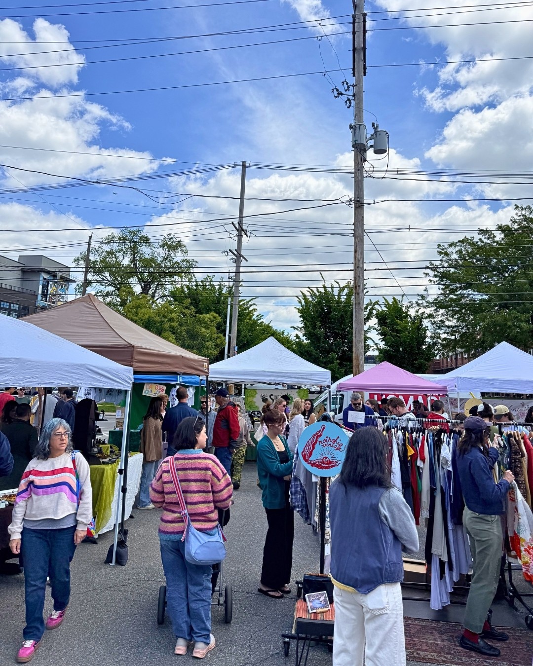 People browse clothing stalls at an outdoor market beneath tents and overhead power lines.