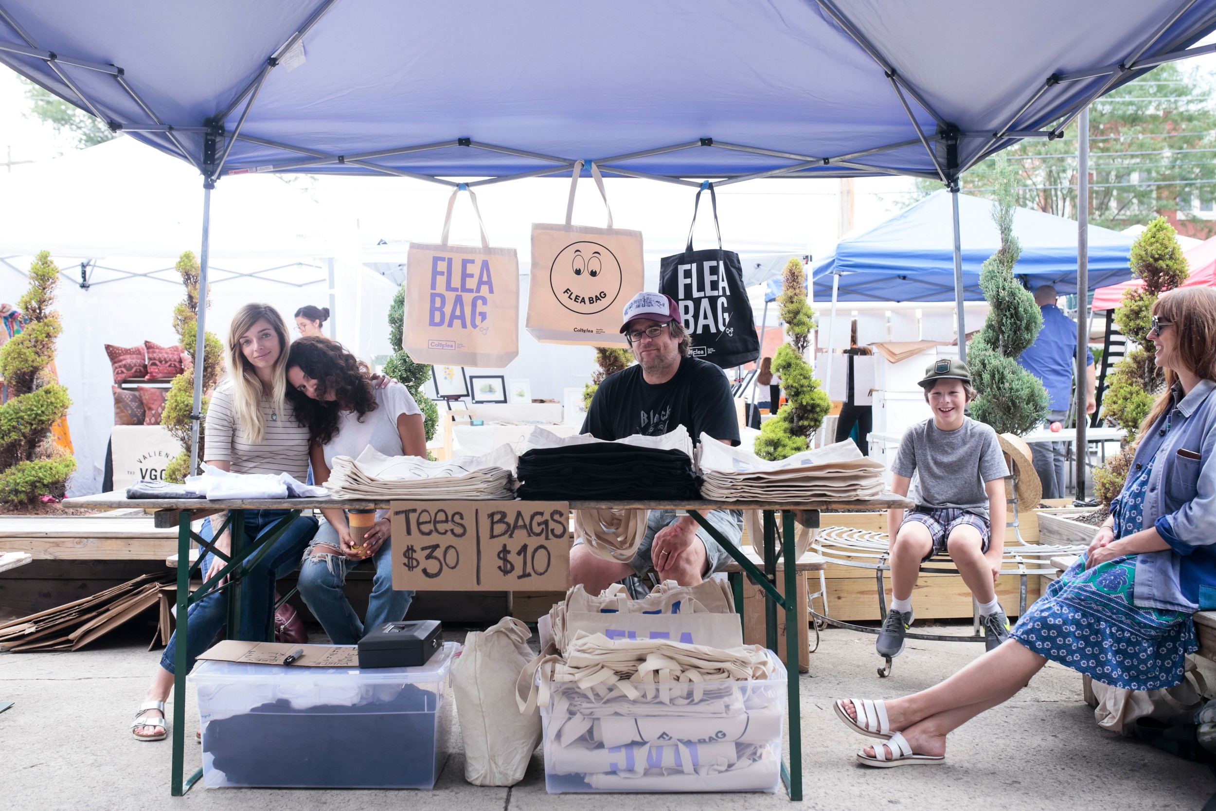 People sit and stand behind a table of baked goods at an outdoor market stall beneath a canopy.
