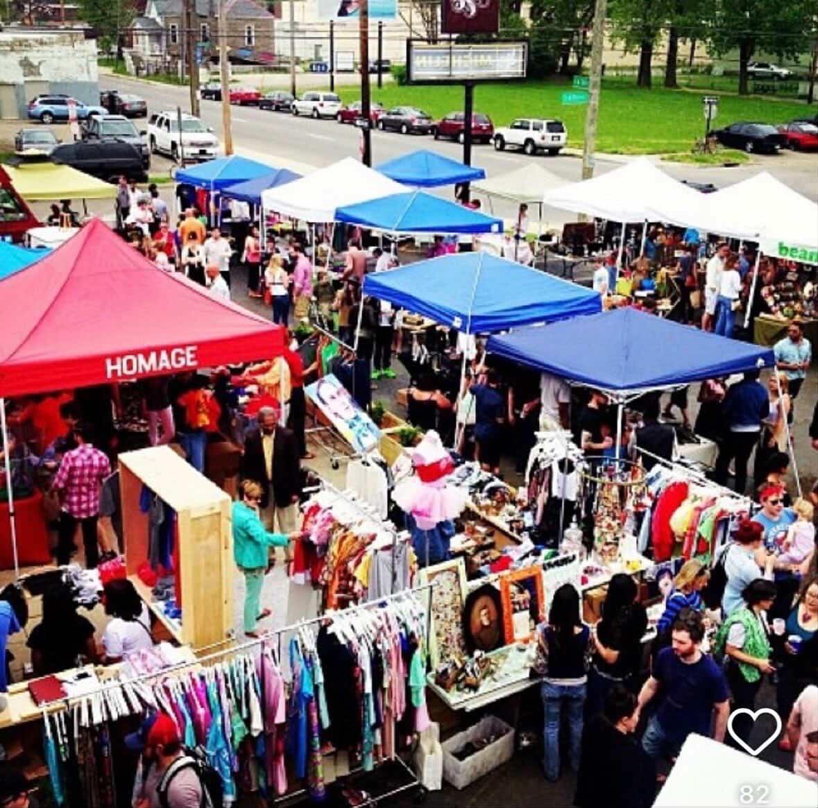 Outdoor market with colorful vendor tents, clothing racks, displays, and people browsing.
