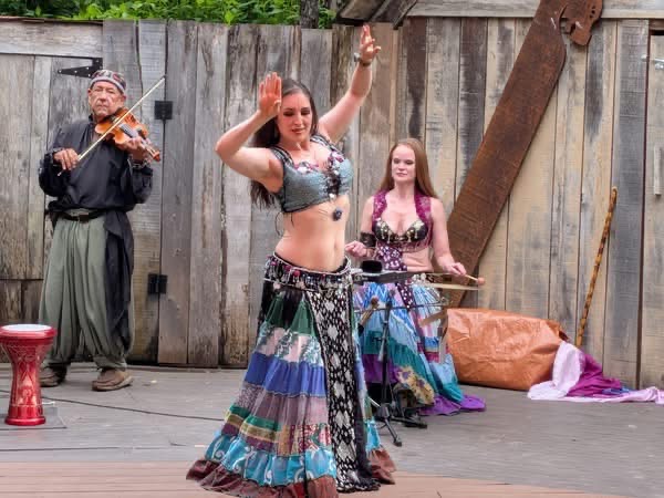 A dancer performs beside a seated musician and a violinist in front of a wooden stage backdrop.