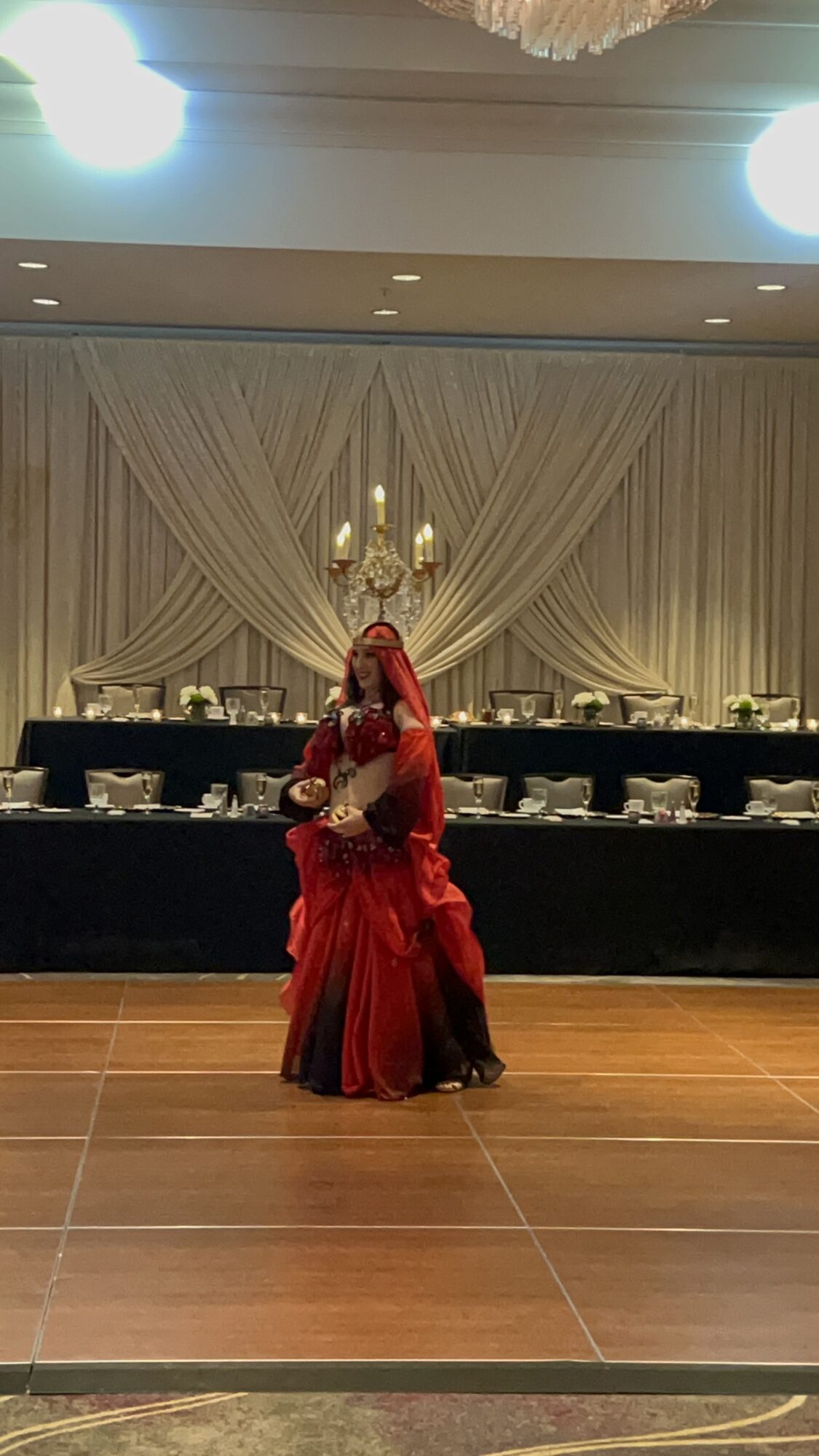 A person in a red gown stands in front of draped curtains and a long table in a ballroom.