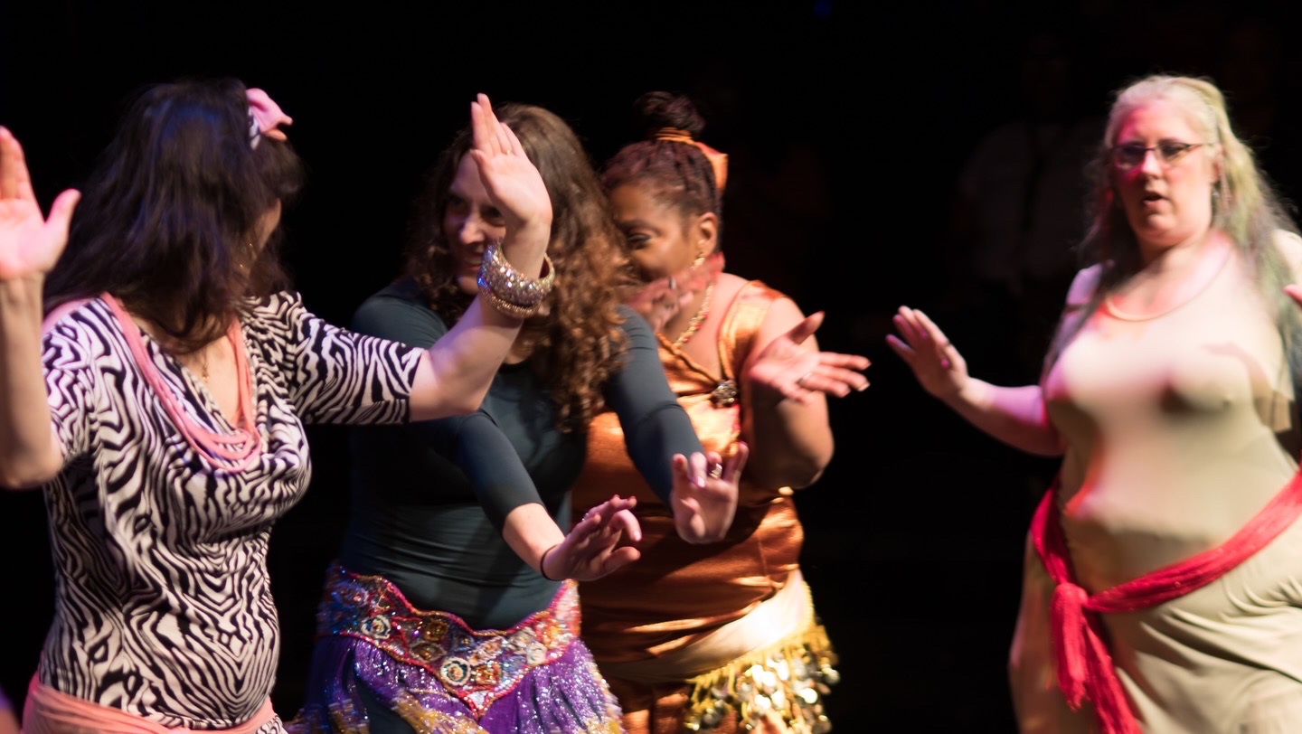 Four people dance on a dimly lit stage, wearing colorful outfits and raising their hands.