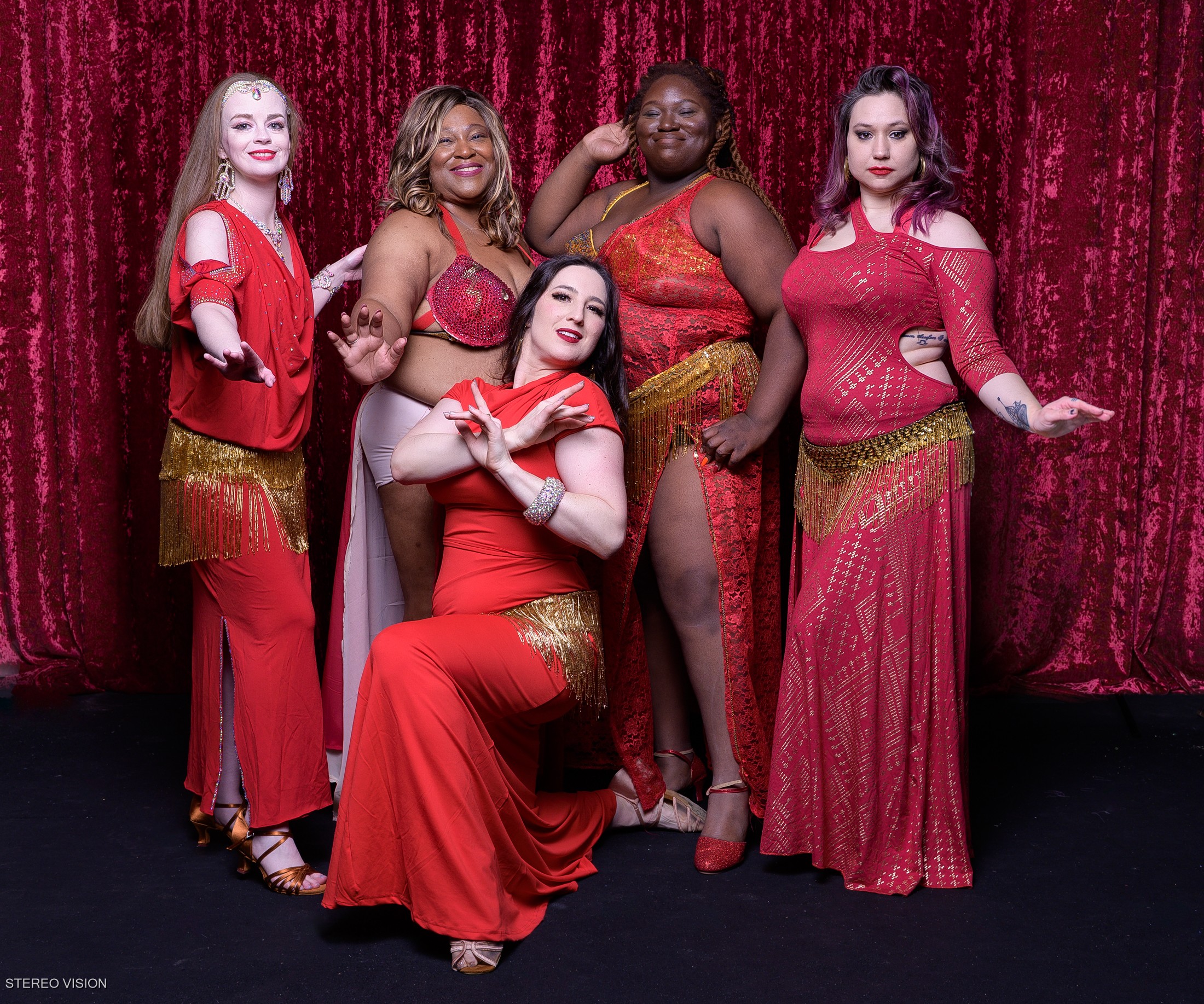 Five people pose in red outfits in front of a red curtain.