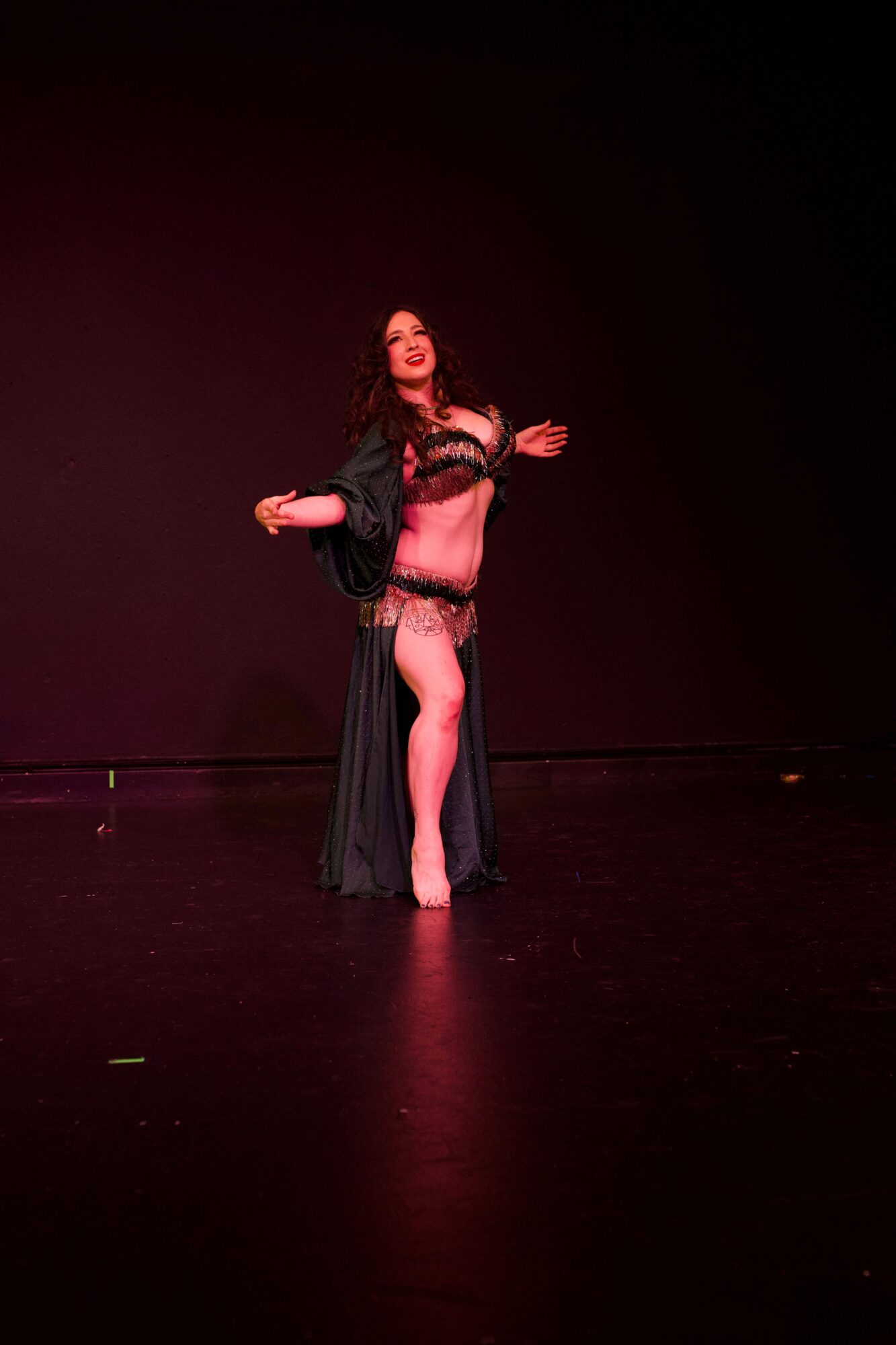 A performer stands on a dimly lit stage in a dark, flowing costume with one leg exposed.