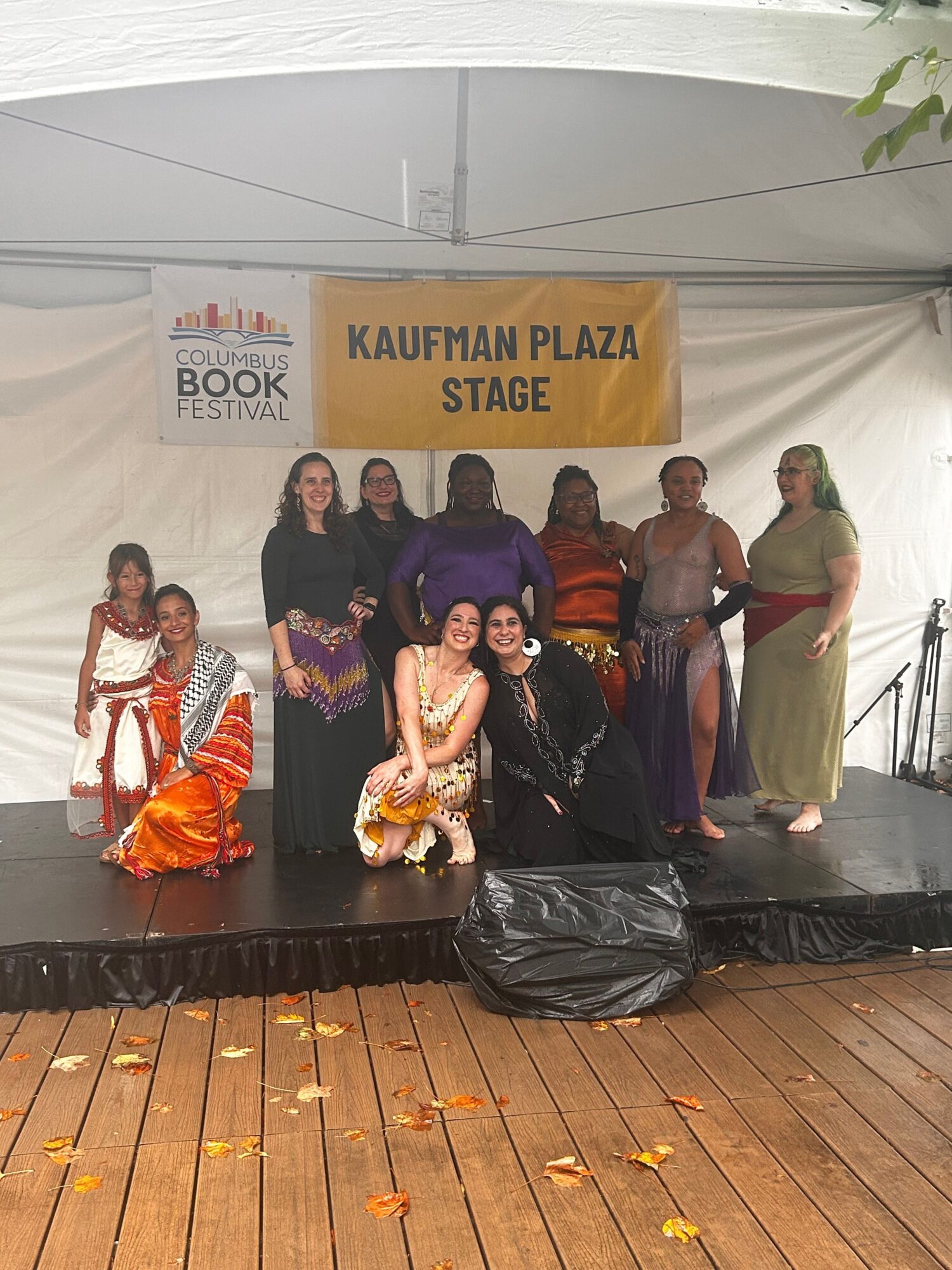 A group of people pose on a stage beside a banner reading “Kaufman Plaza Stage.”