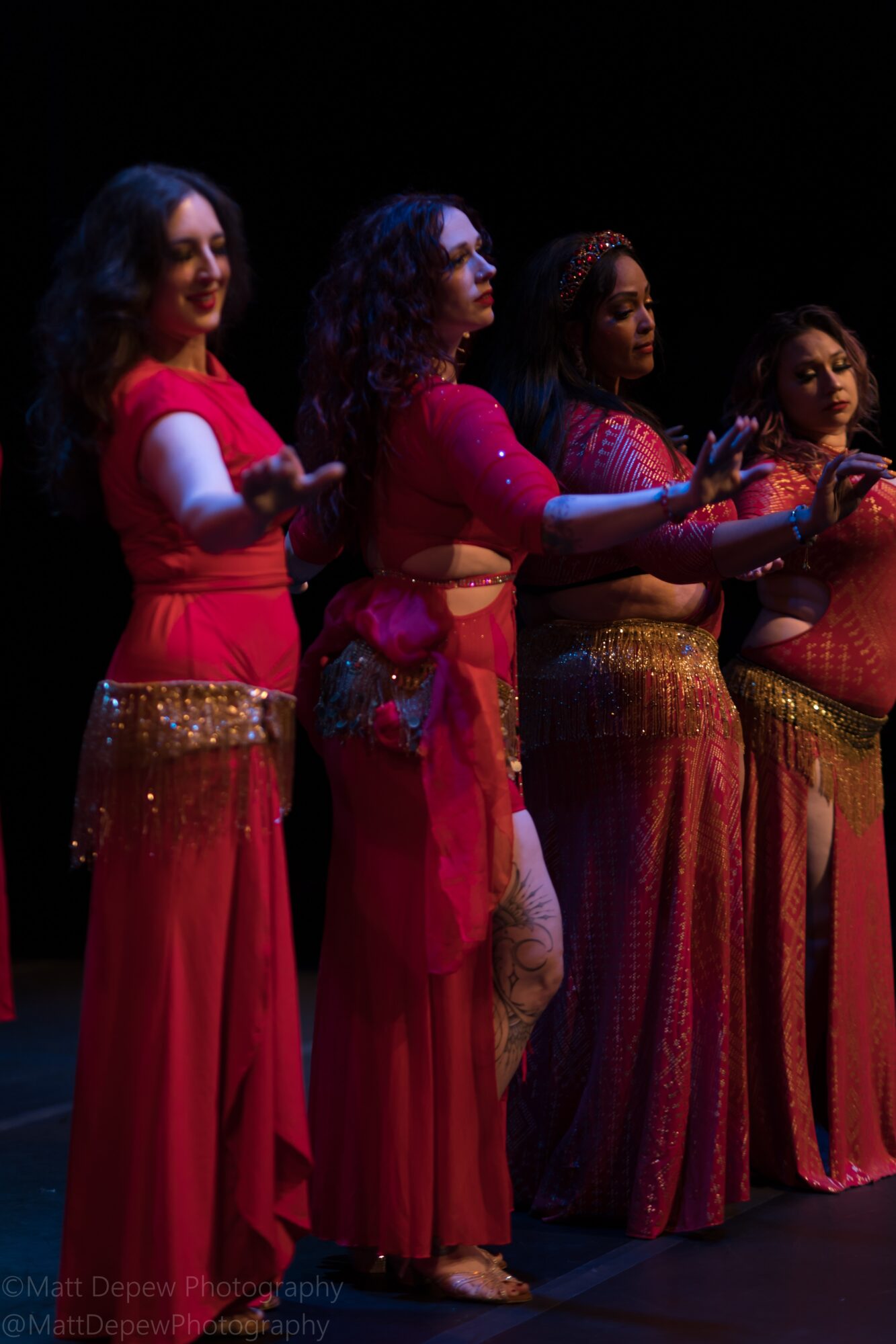Four performers in long, colorful dresses stand in a row under stage lighting.