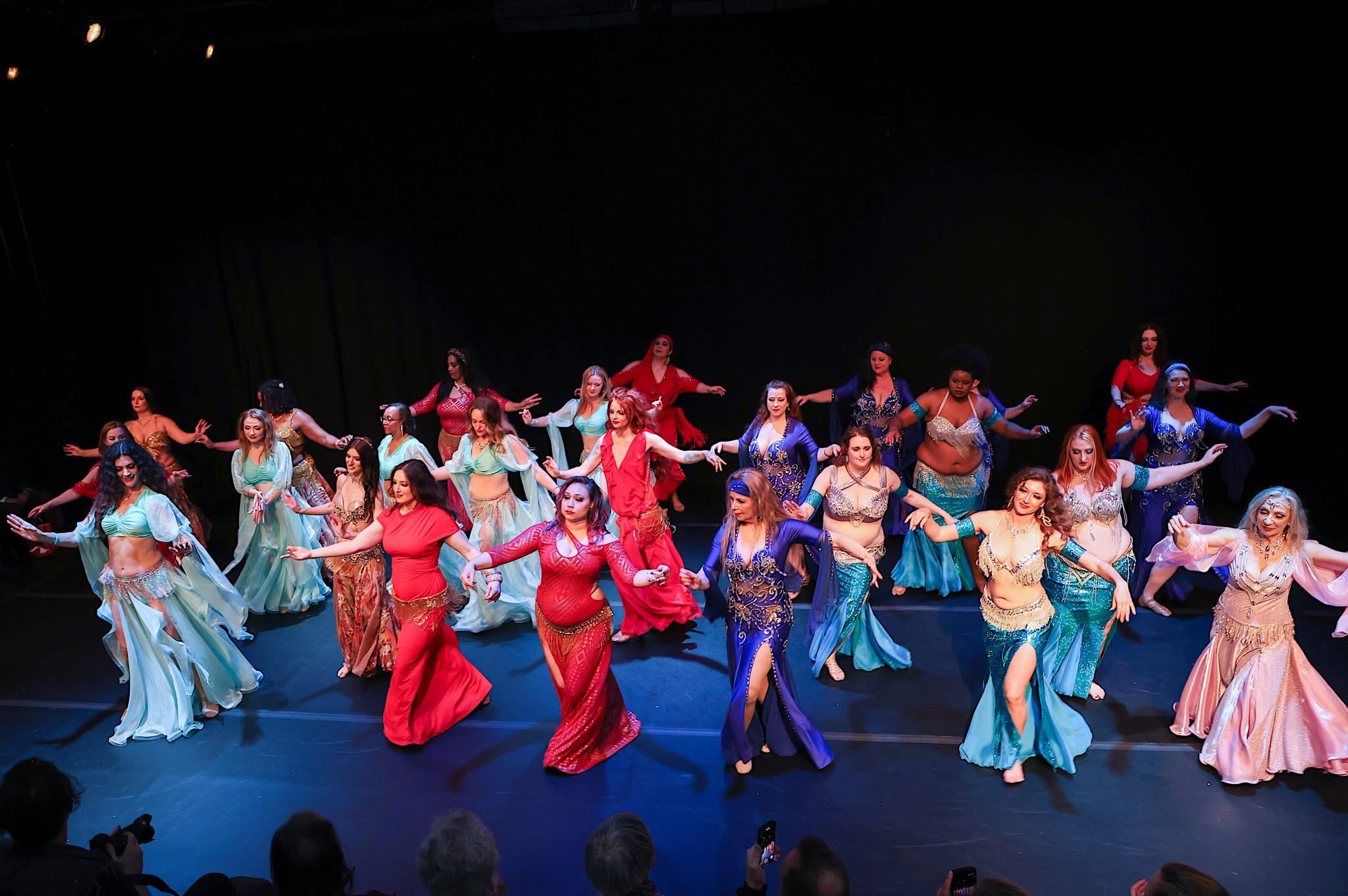 A large group of performers in colorful costumes dance across a theater stage.