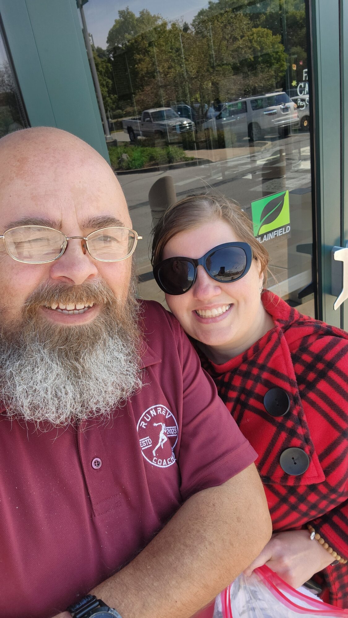 Two people pose for a selfie outside a glass storefront, with trees and parked vehicles reflected in the window.