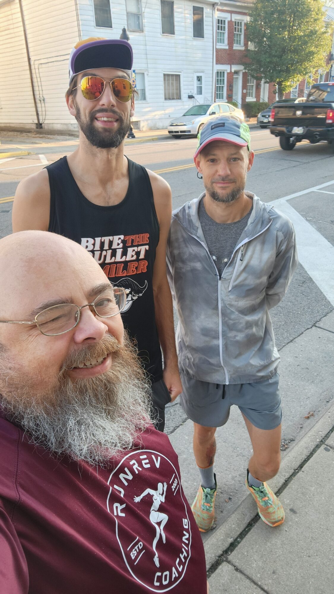 Three people pose together on a sidewalk beside parked vehicles and residential buildings.
