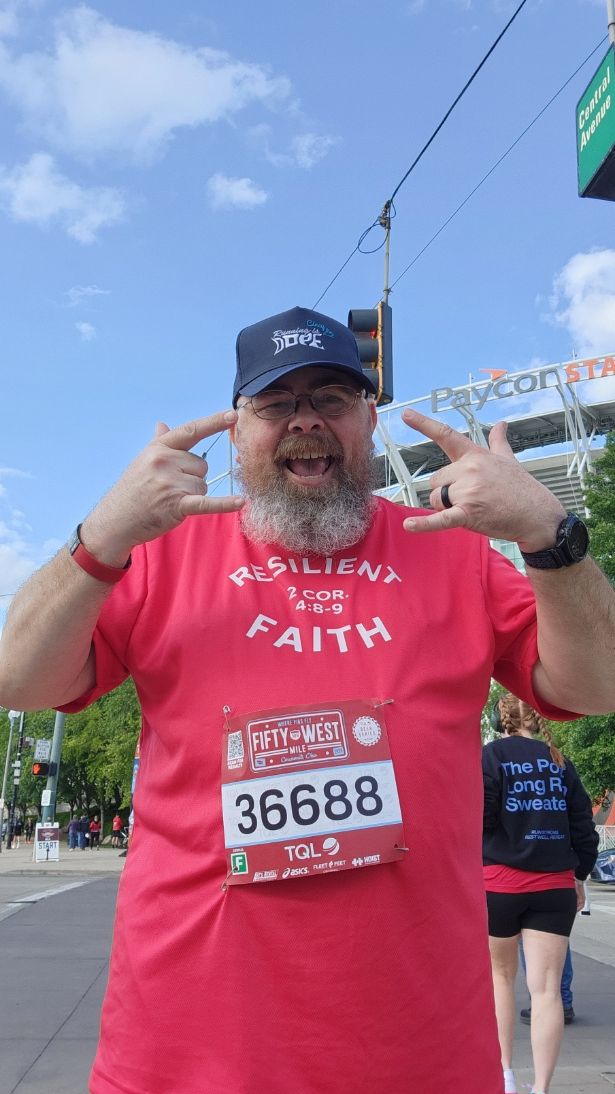 A person in a red shirt and blue cap poses beside a race route under a clear sky.
