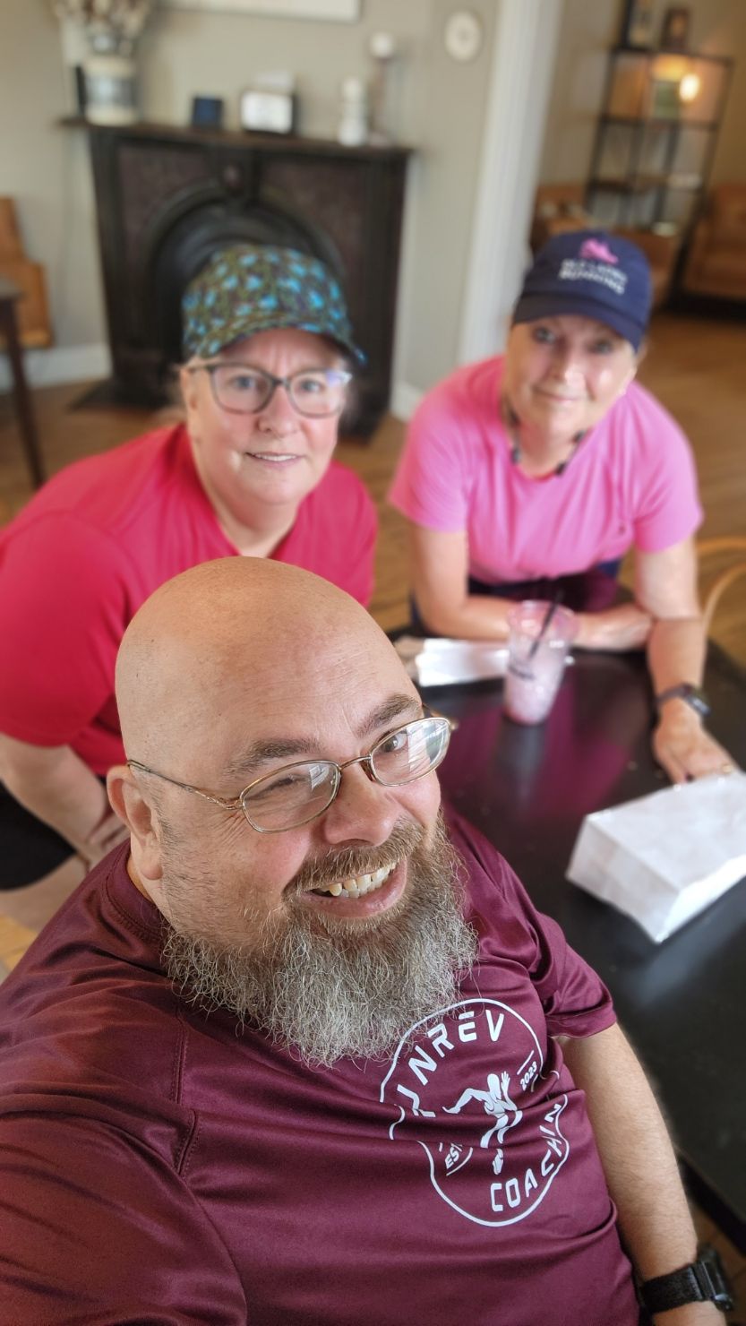 A seated bearded person in a maroon shirt poses with two people wearing pink shirts in a room.