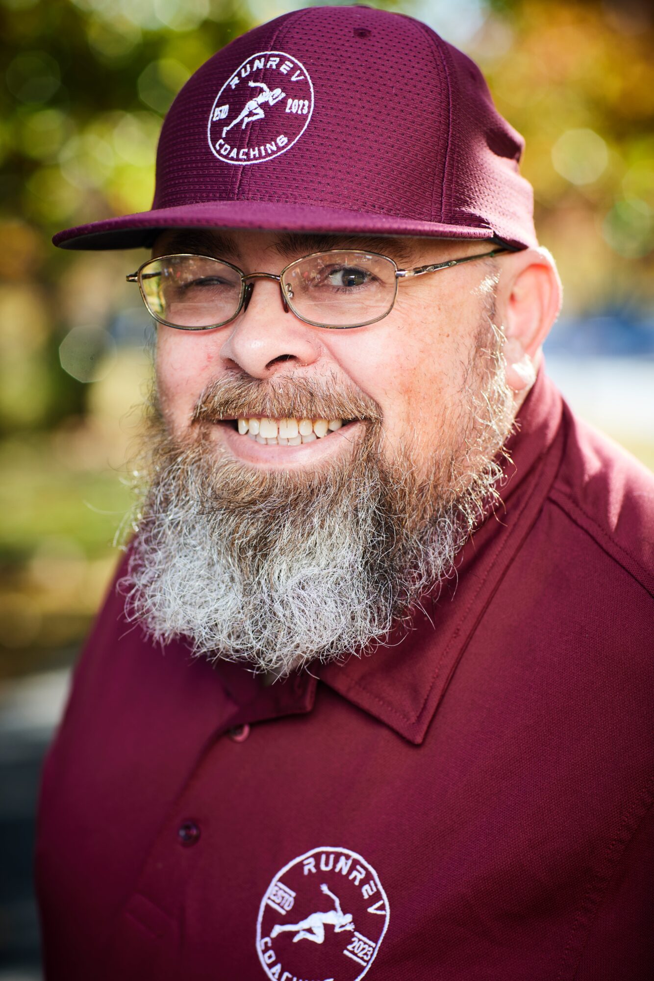 Person wearing a burgundy cap and shirt, glasses, and a long gray beard, with greenery in the background.