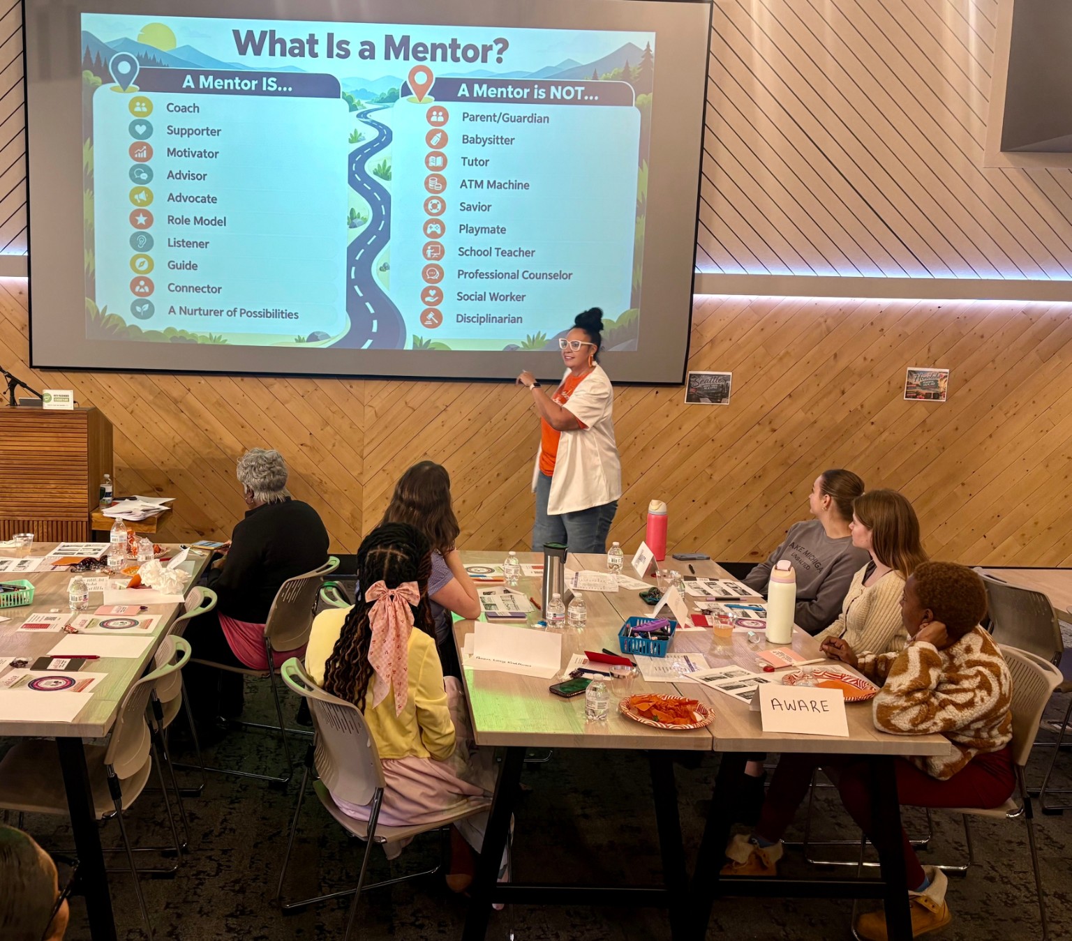 A presenter points to a projected mentor infographic while seated attendees watch around tables.