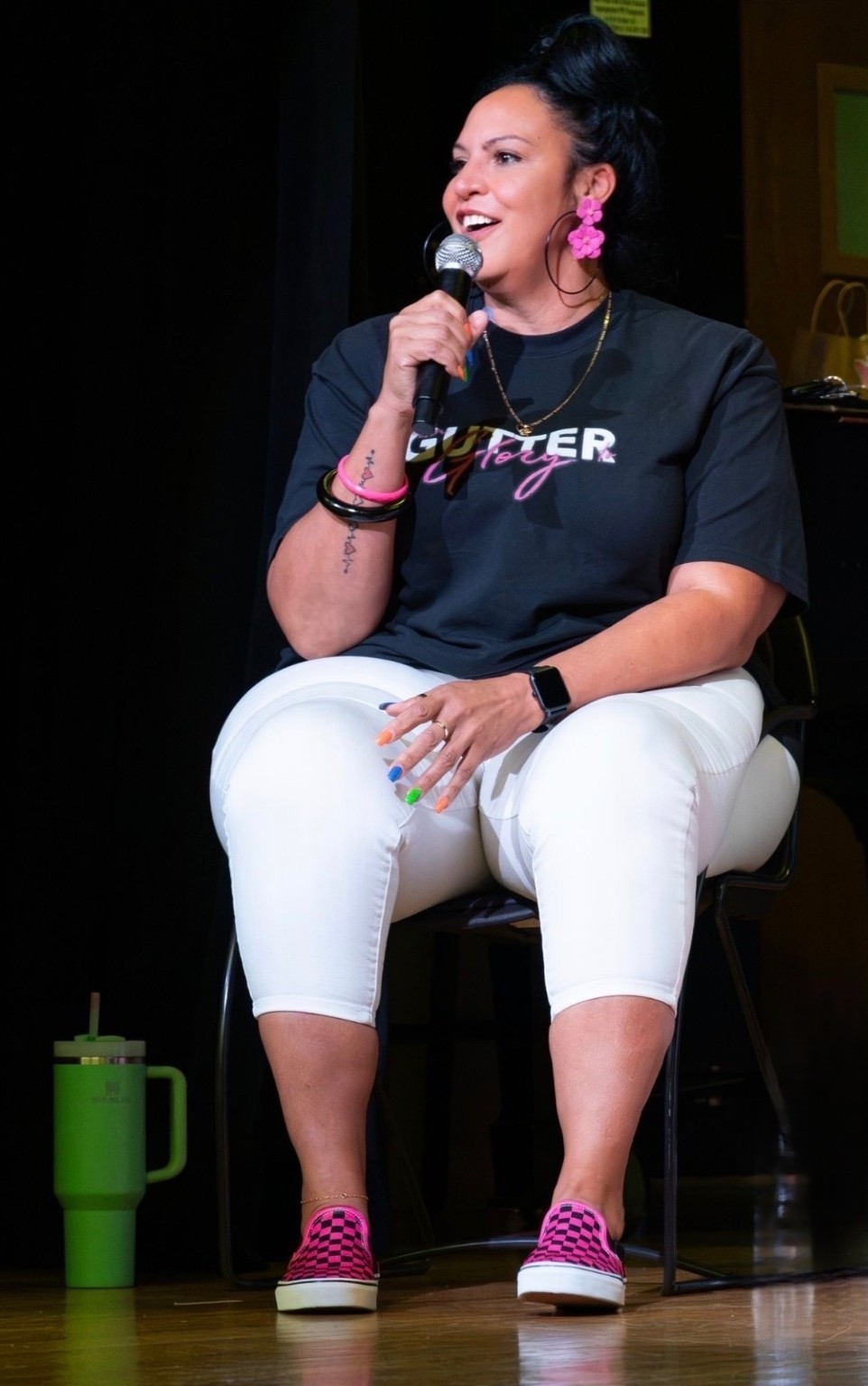 A person sits onstage holding a microphone, wearing a black shirt, white pants, and bright pink shoes.