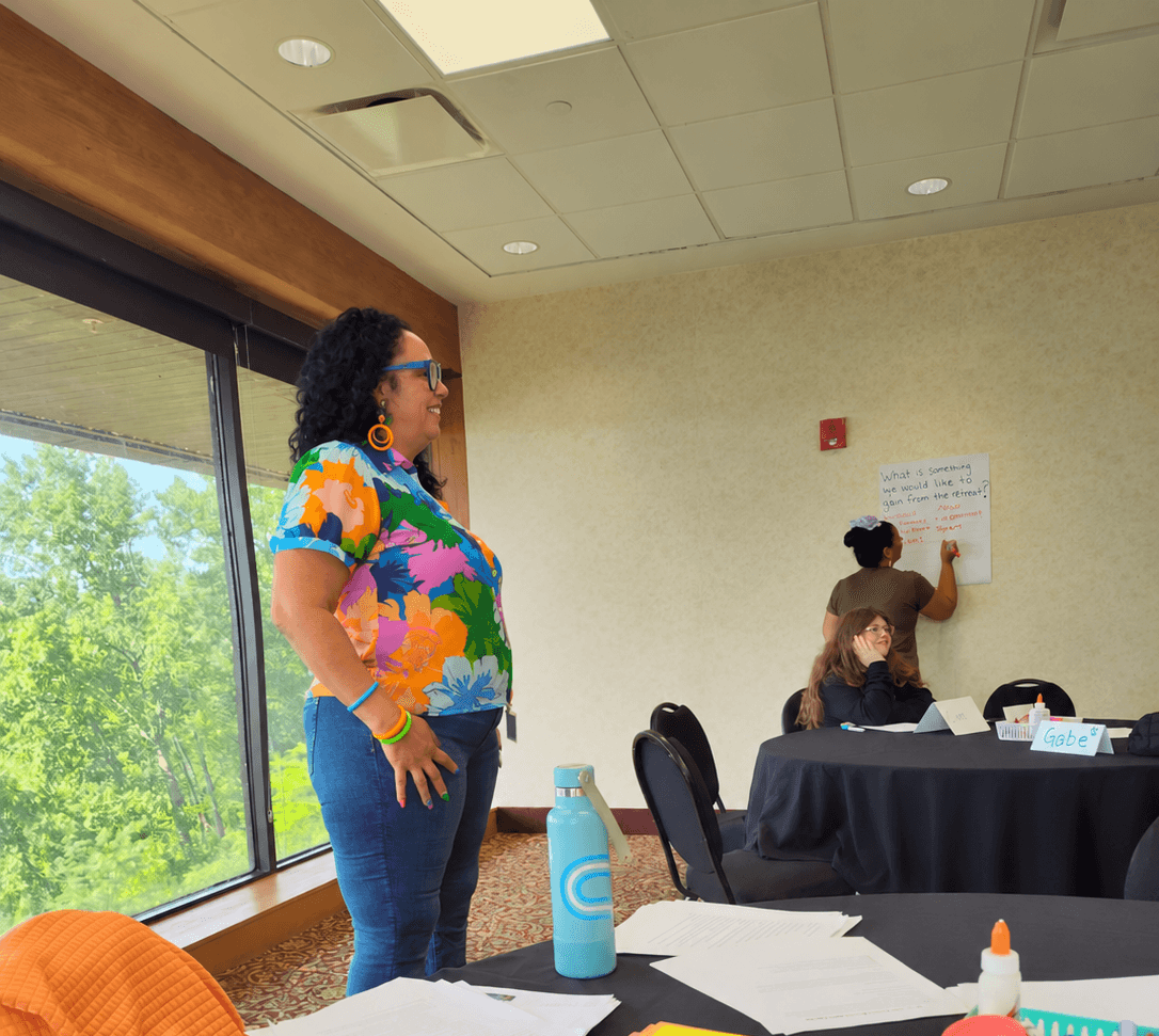 A person in a colorful shirt stands beside a window while two people sit at a table in a meeting room.