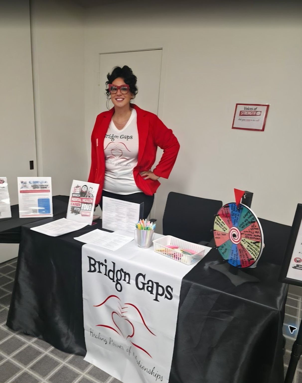 Person standing behind a table displaying Bridge Gaps materials, pamphlets, and a colorful prize wheel.