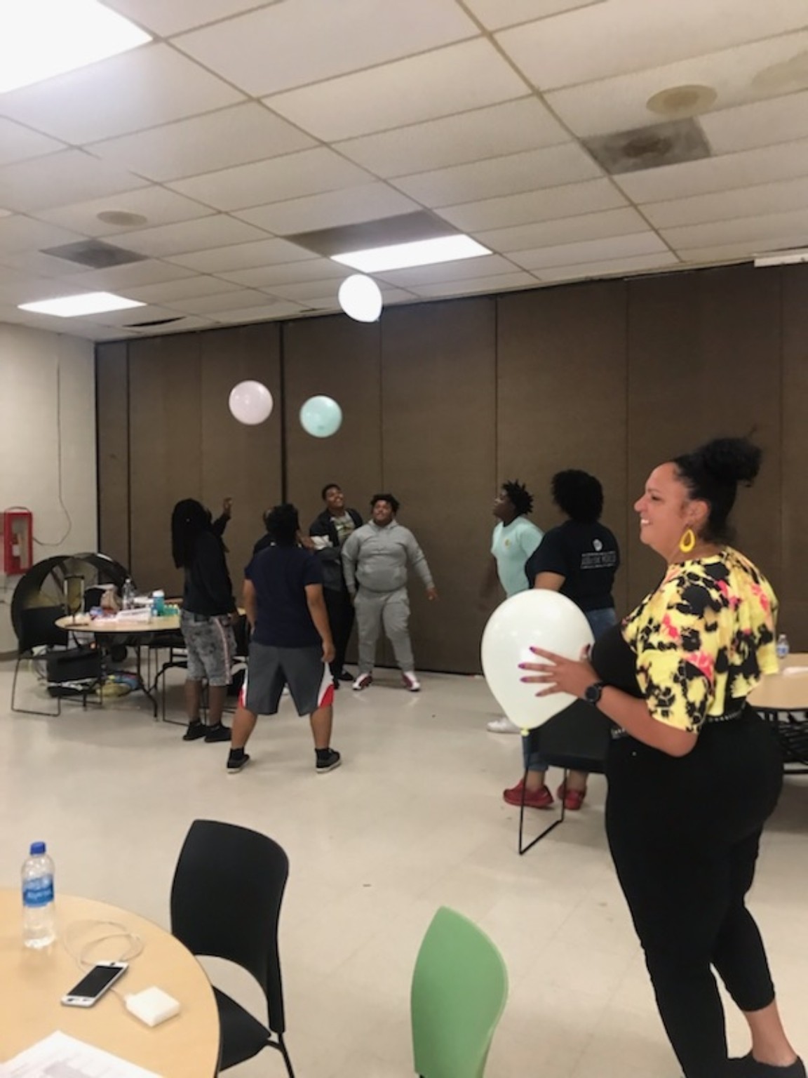 People gather in a room with balloons, tables, chairs, and a person holding a large white balloon.