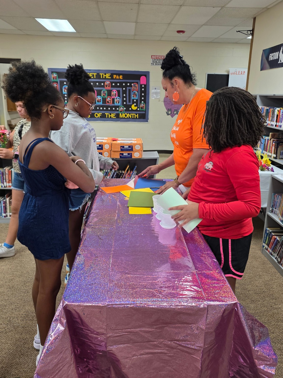 Three people gather around a purple-covered table with small containers and papers in a classroom.