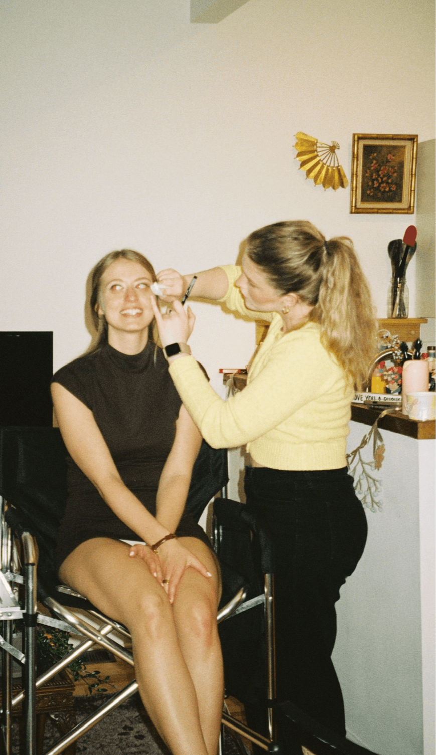 A seated person has makeup applied by another person in a yellow top.