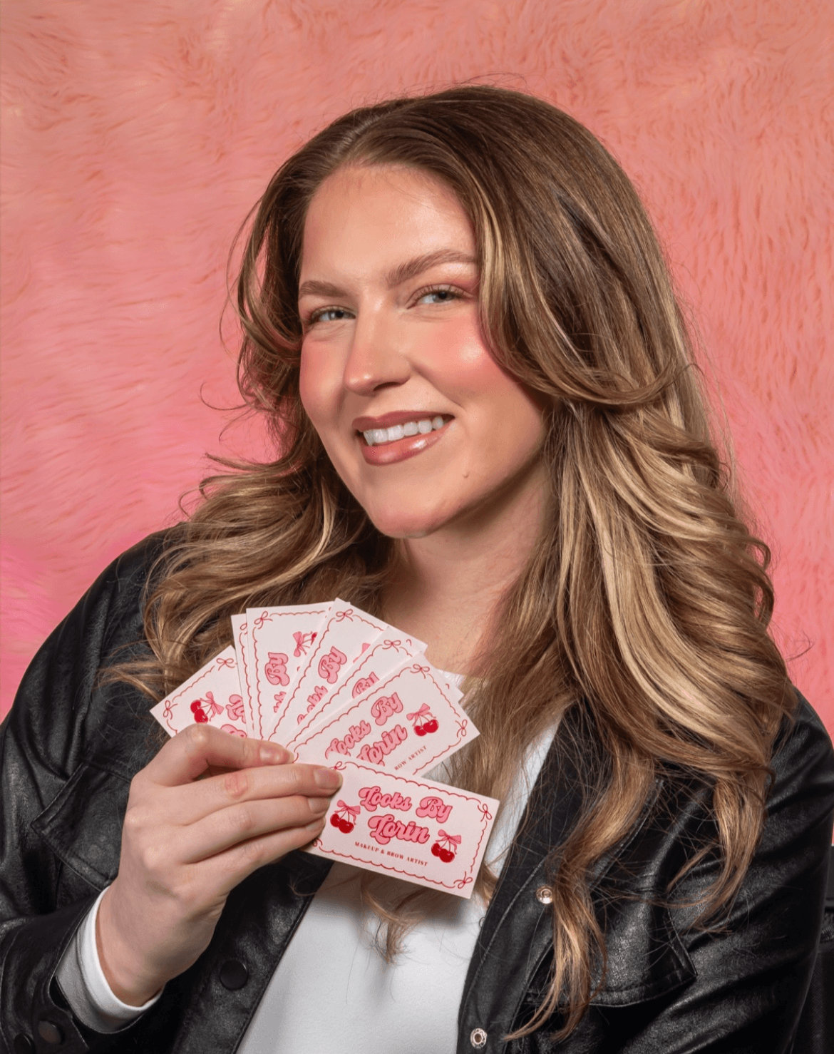 A person with long wavy hair holds several pink playing cards against a textured pink backdrop.