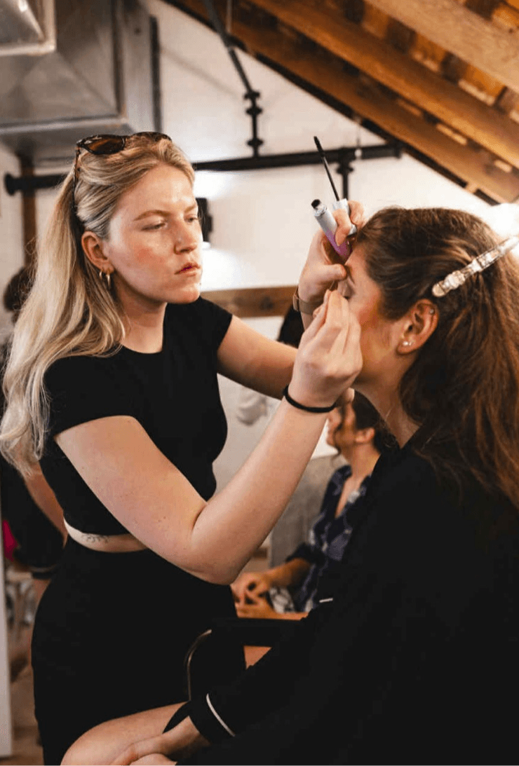 A makeup artist applies eye makeup to a seated person beneath exposed wooden ceiling beams.