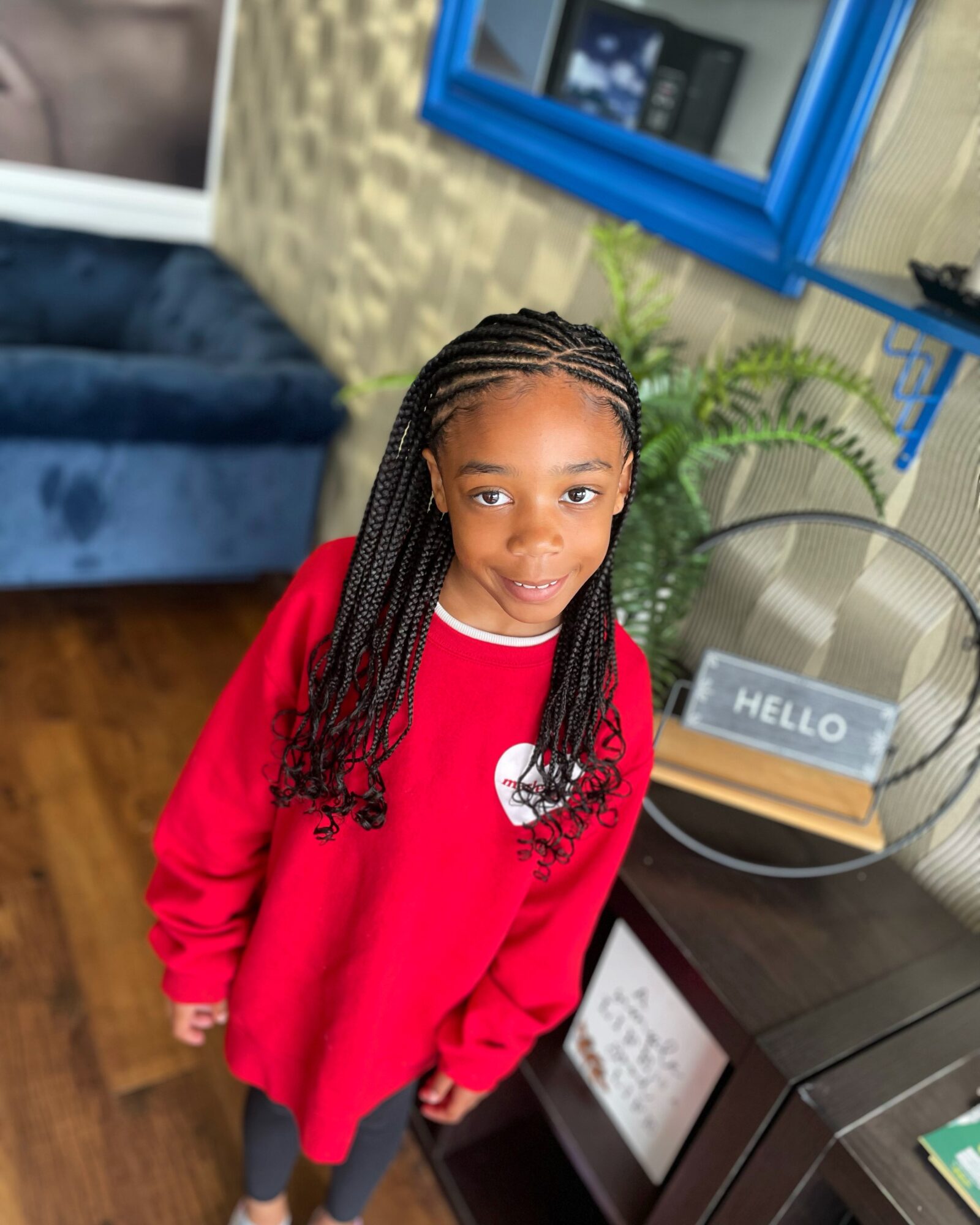 A child with braided hair stands in a red sweatshirt beside a chair and table in a living room.