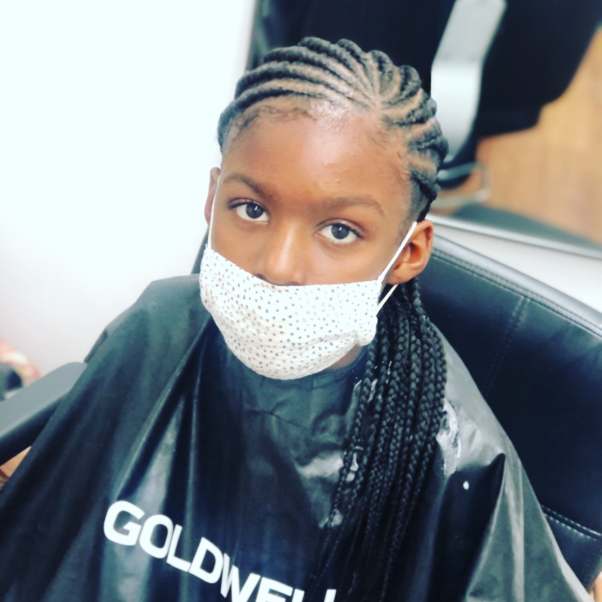 Person with braided hair wearing a face mask and a black salon cape.