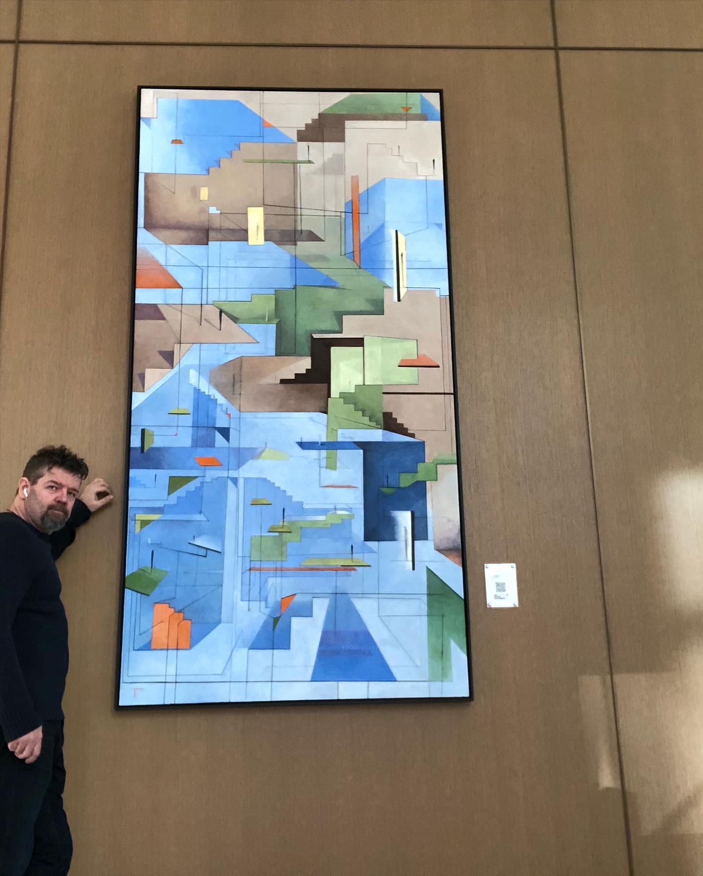 A person stands beside a tall framed geometric artwork mounted on a brown wall.