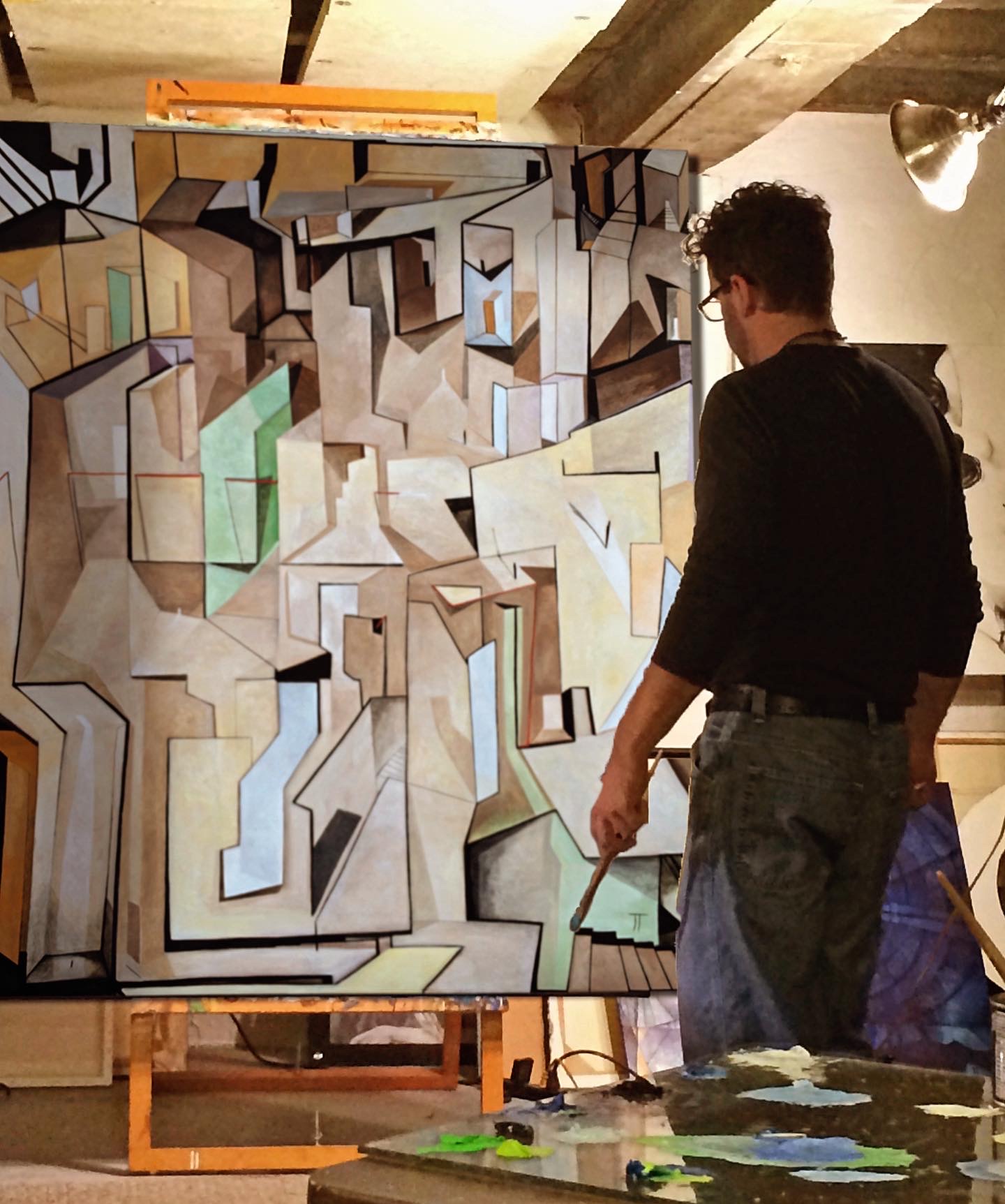 A person stands beside a large geometric mural in a studio, holding a paintbrush near its lower edge.