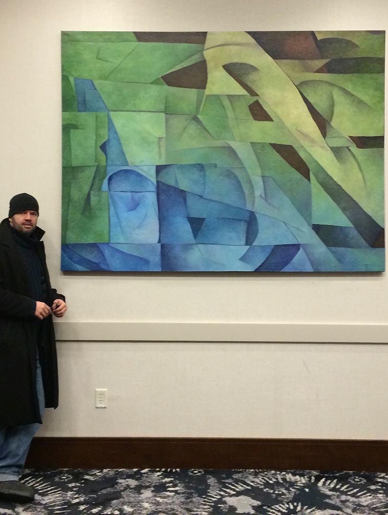 A person stands beside a large abstract painting with green and blue geometric shapes on a gallery wall.