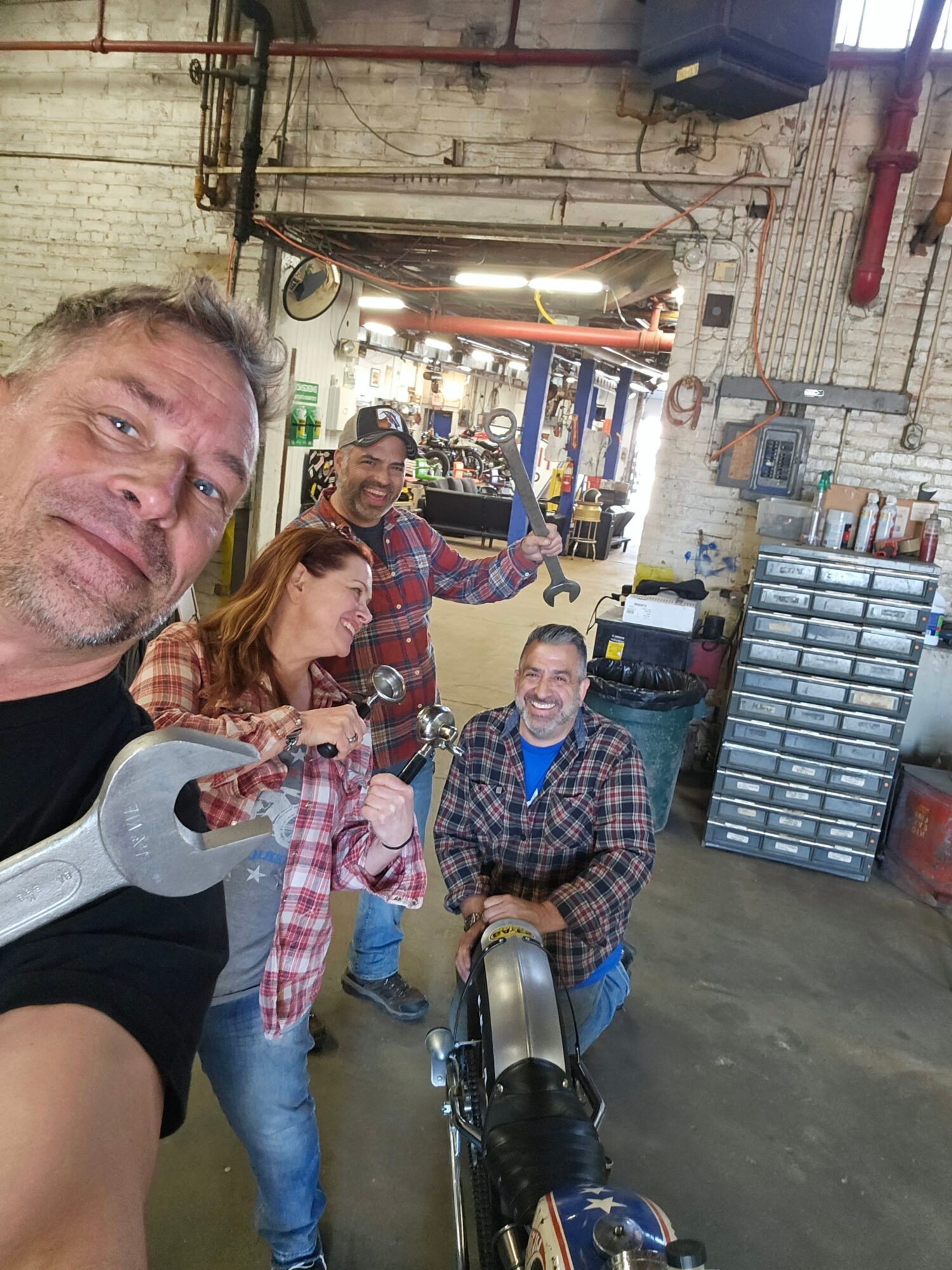 Three people pose in a workshop, with a large wrench held close to the camera.
