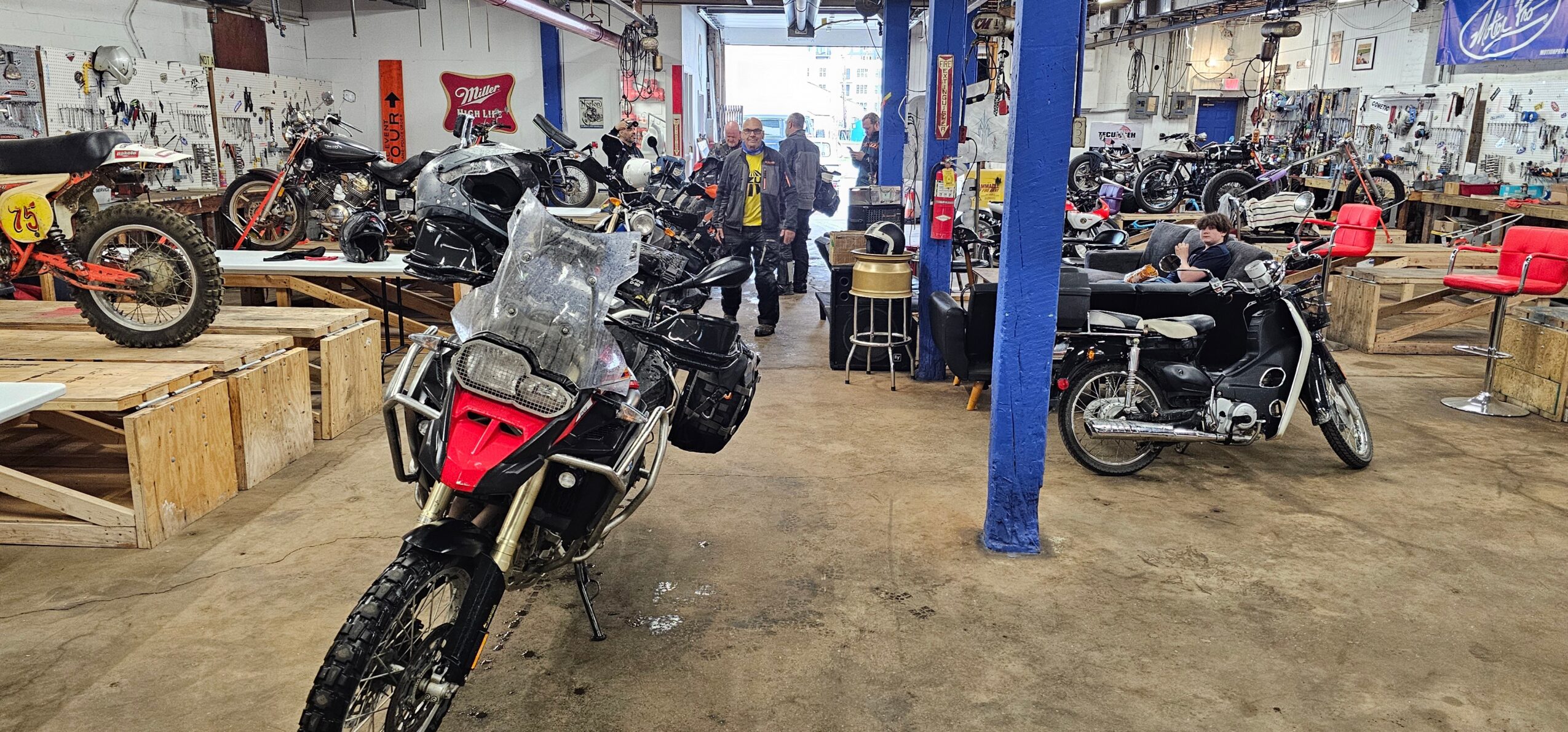 Motorcycles and people inside a workshop with blue support columns and tools.