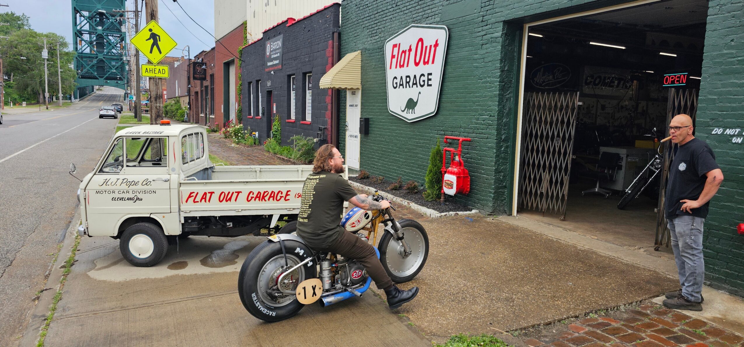 A person sits on a motorcycle outside a garage, with a small service truck and another person nearby.