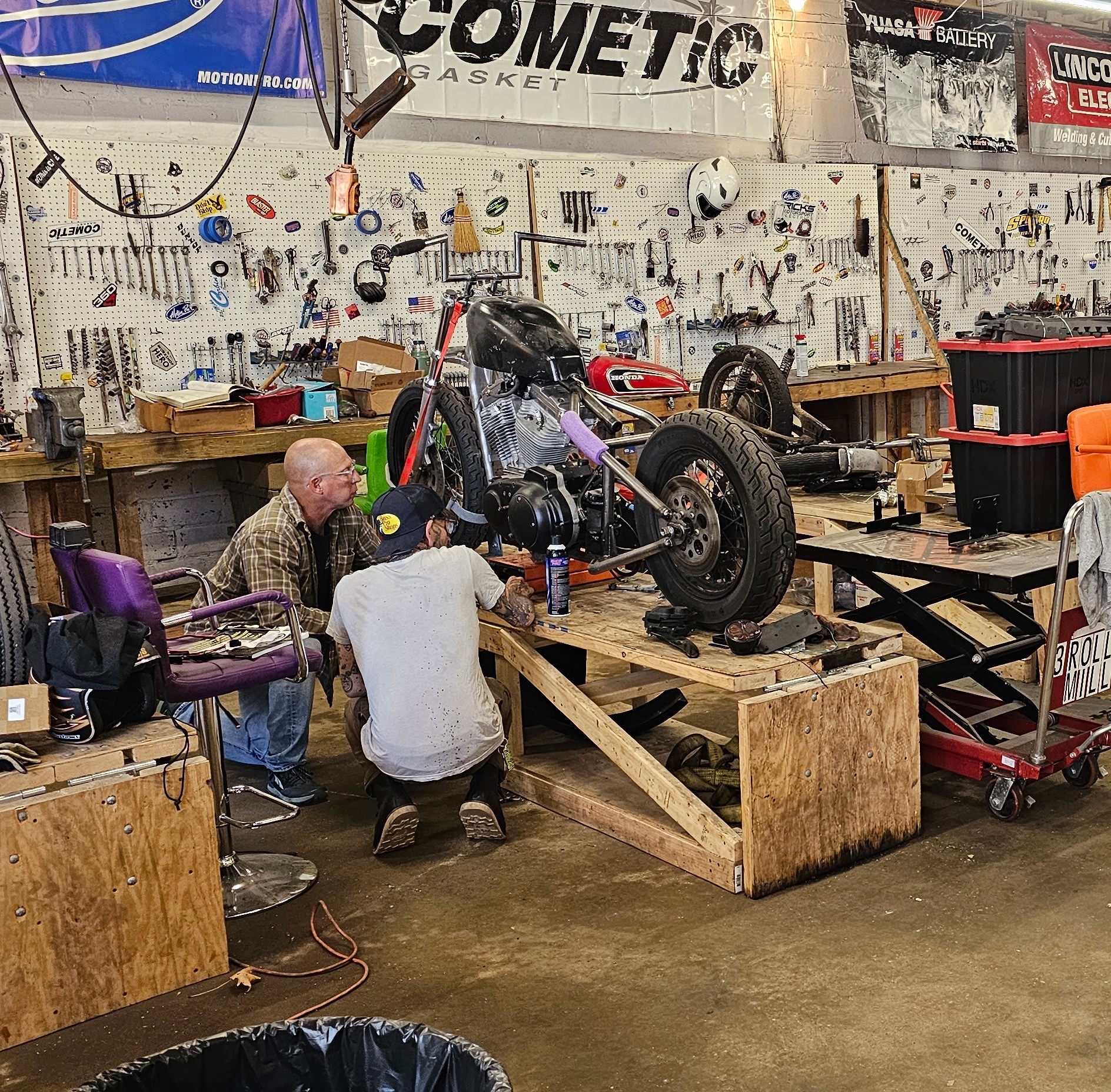Two people work on a racing motorcycle mounted on a wooden platform in a workshop filled with tools and equipment.