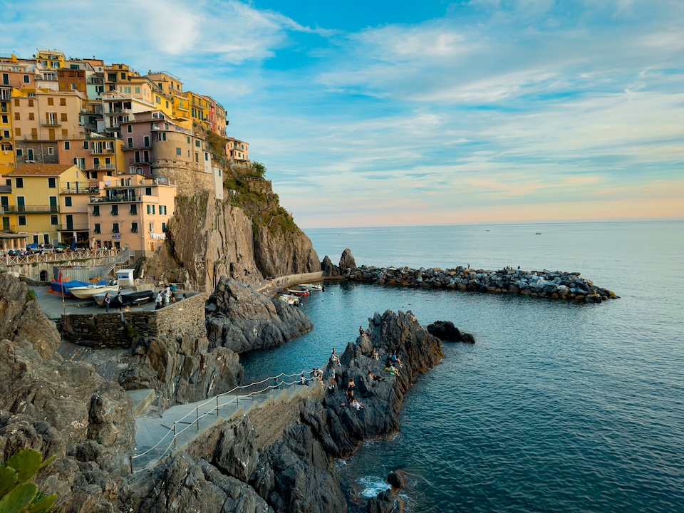 Colorful buildings on a rocky coastal cliff overlook a small harbor and calm sea.