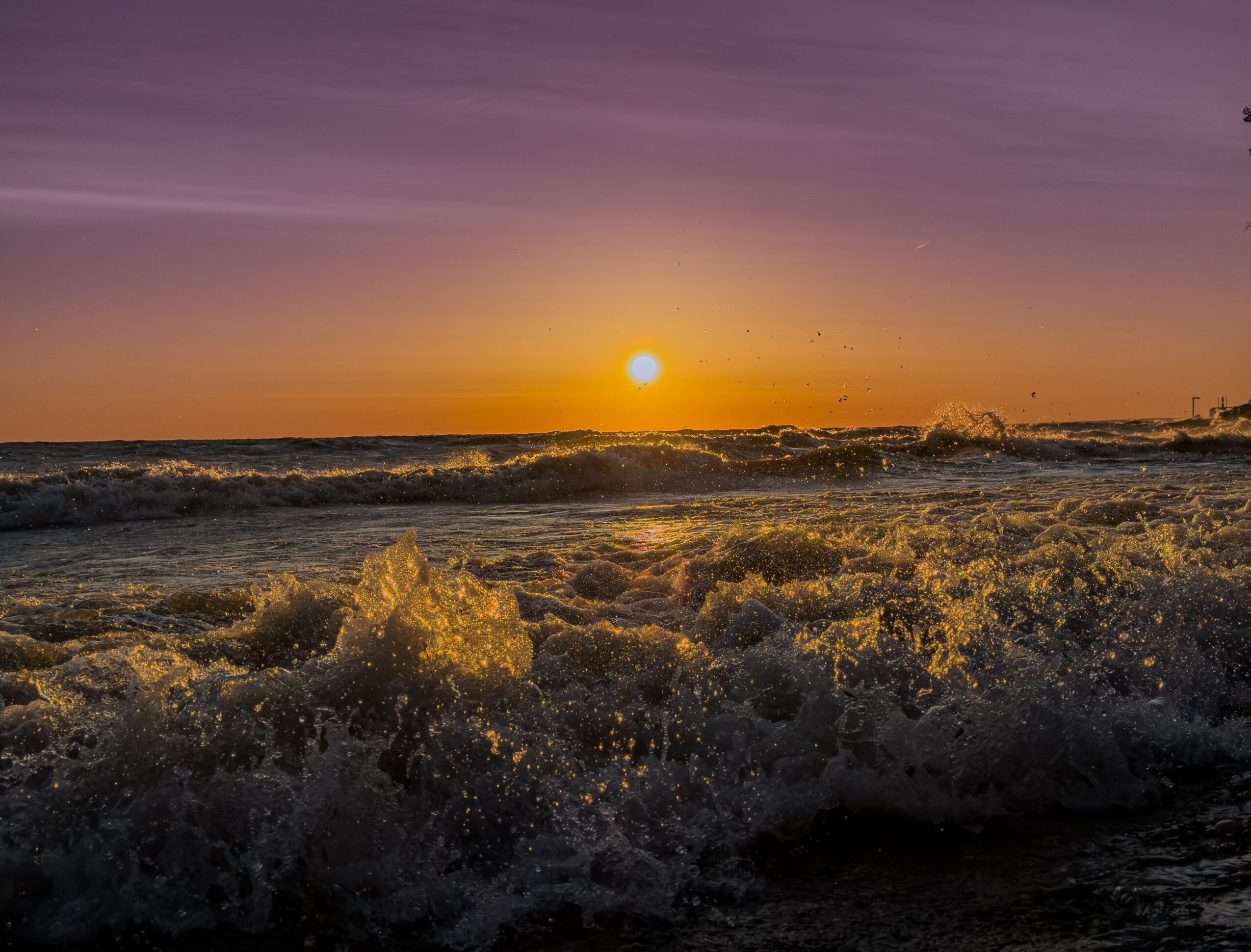 Sunset over the ocean, with waves breaking in the foreground and a purple-orange sky.