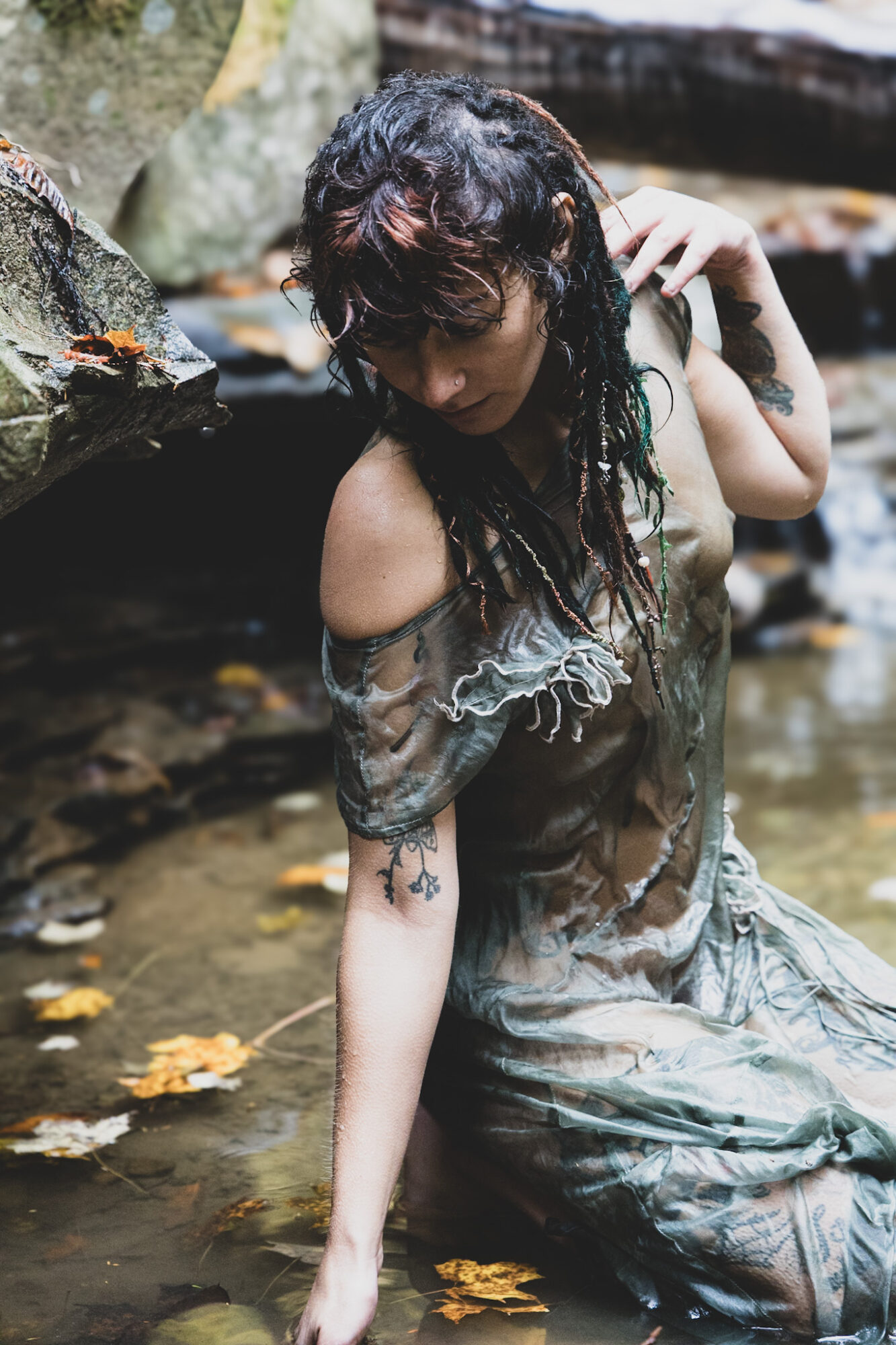 Person in a wet, flowing outfit kneels in a shallow stream, with one hand near their head.