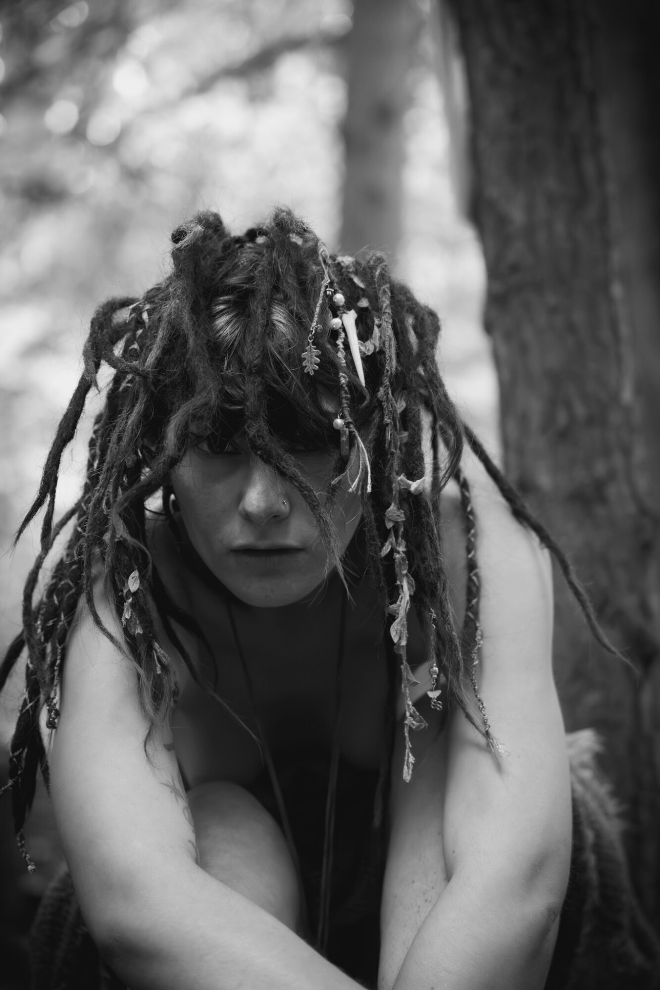A person with long, decorated dreadlocks crouches in front of a tree in a wooded area.