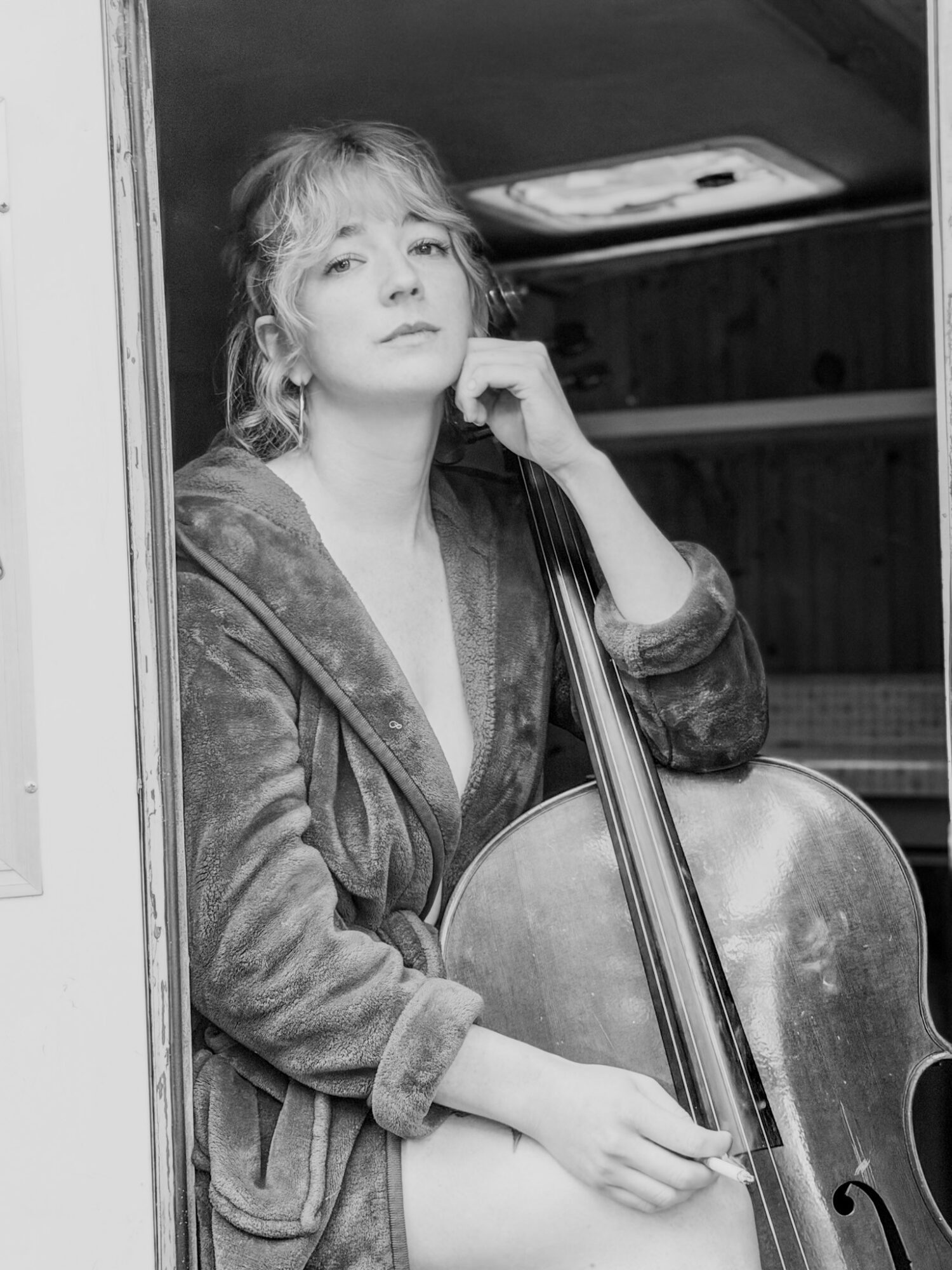 A person seated in a doorway holds a cello, with one hand resting near their face.