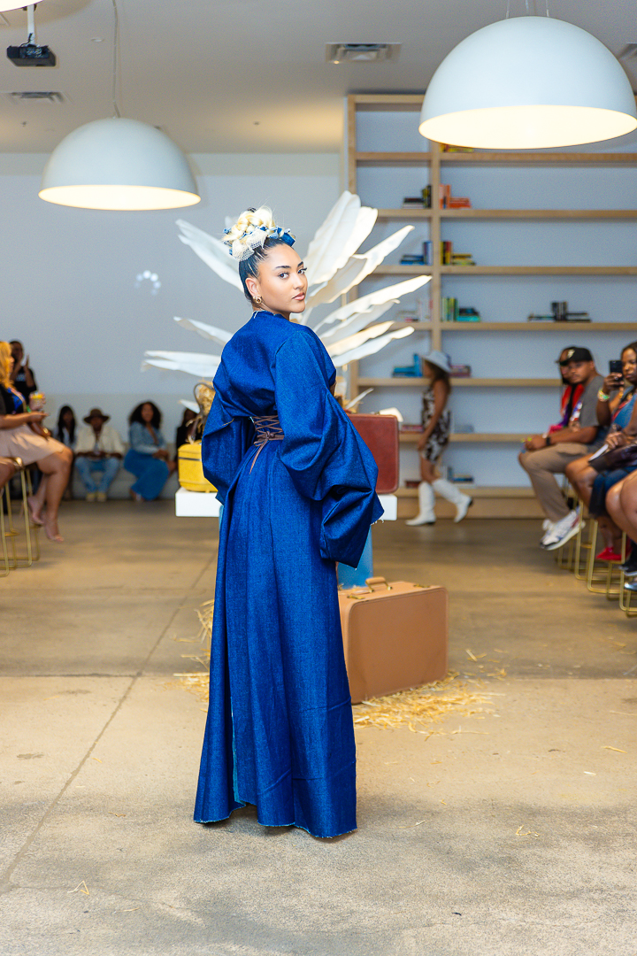 A person in a long blue garment and white feathered headpiece stands in a room with seated spectators and hanging lights.