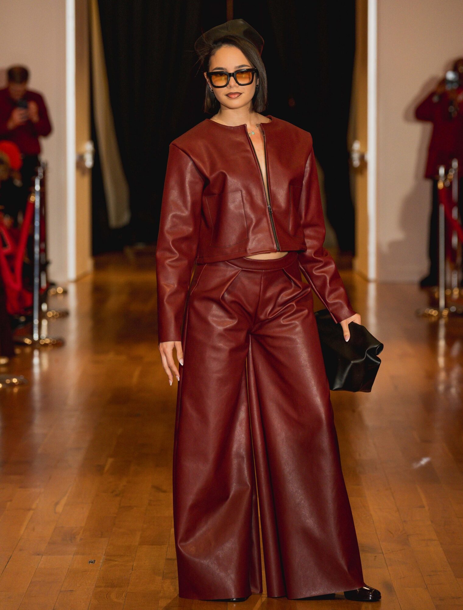 Person wearing a red leather jacket and wide-leg trousers walks down a runway carrying a black handbag.